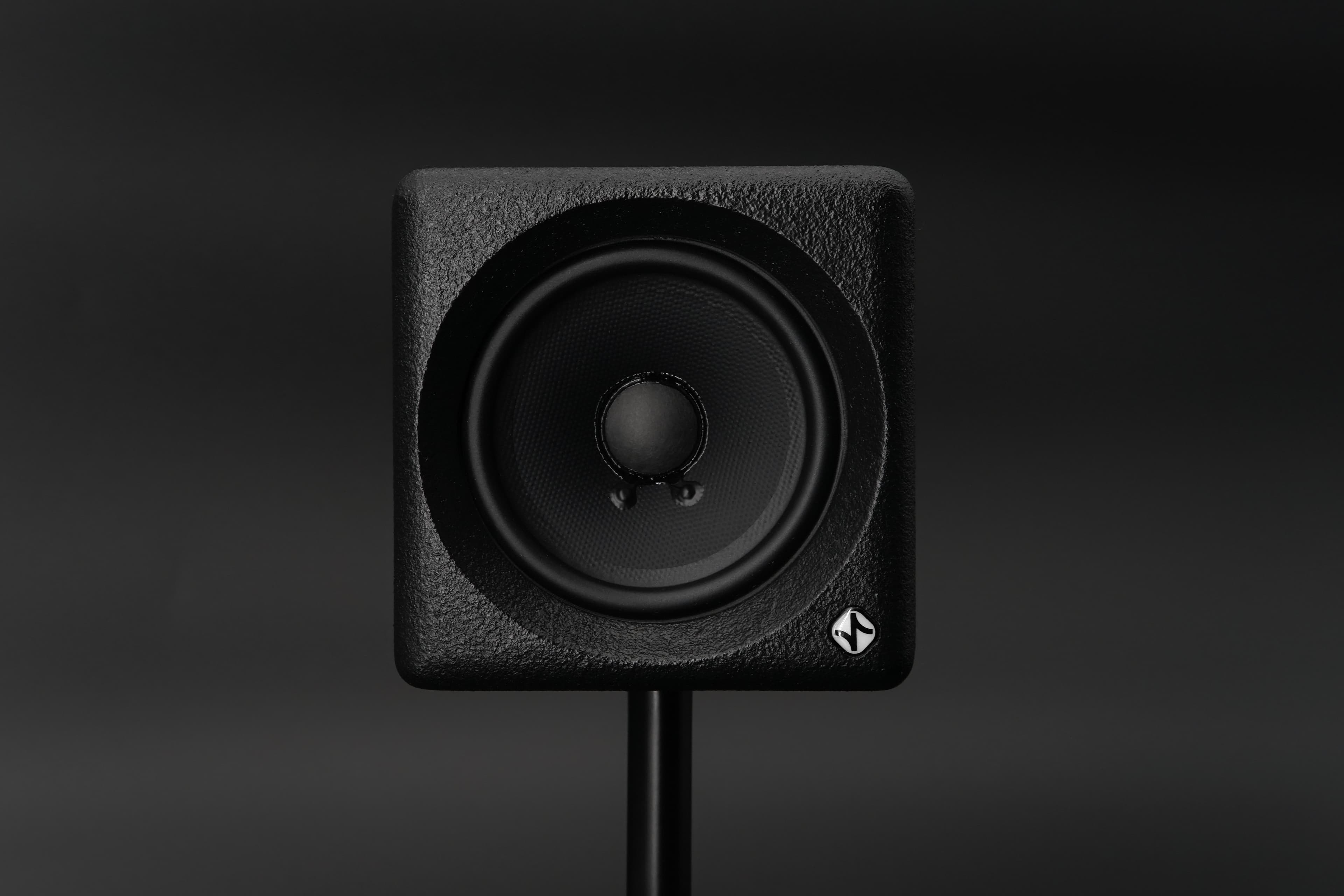 A small black cube studio monitor with a single full-range driver, on a stand against a dark background.