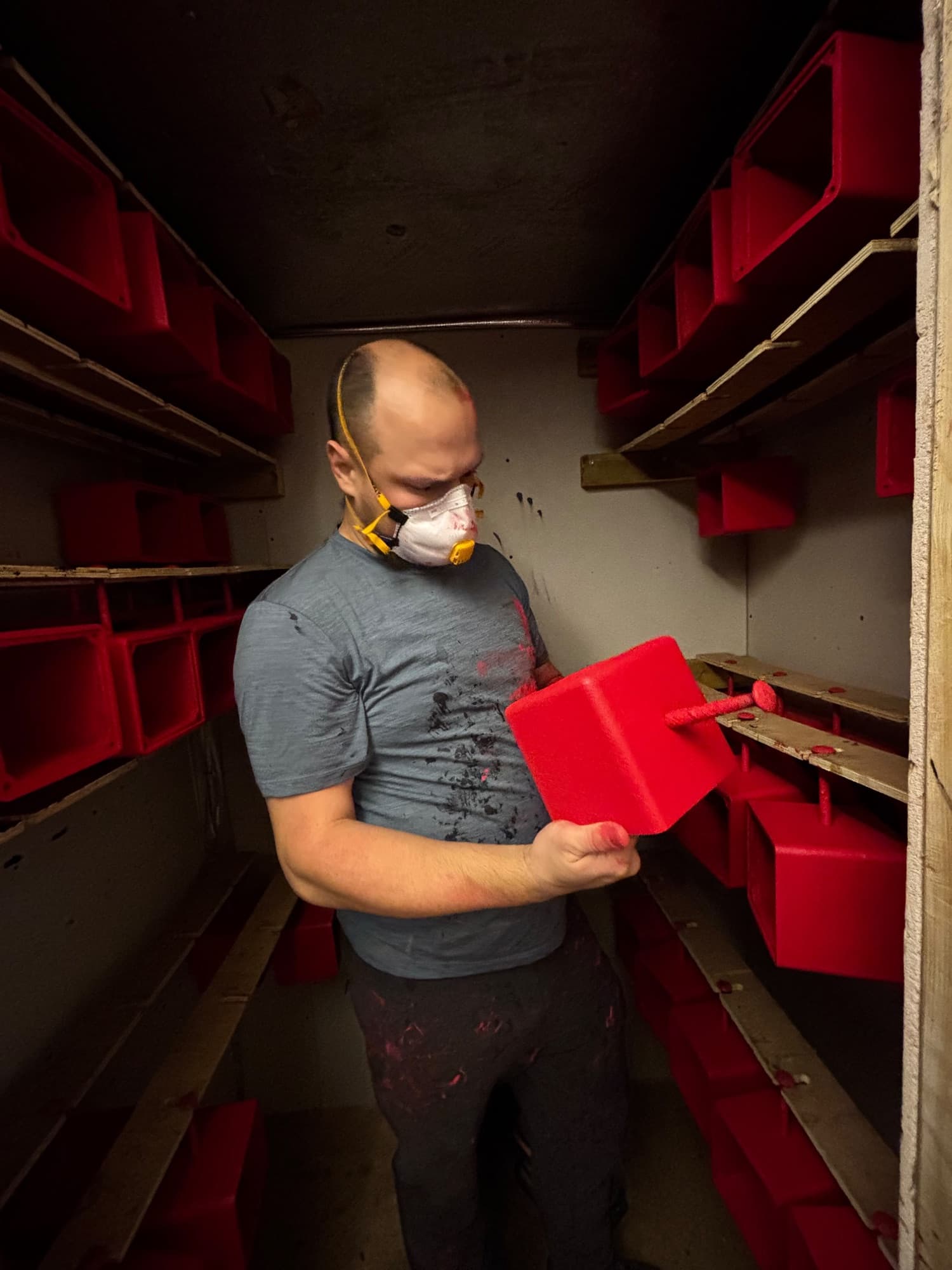A Tantrum Audio builder in a dust mask checking a freshly sprayed red Angry Box enclosure in the spray booth, with racks of painted enclosures curing on both sides