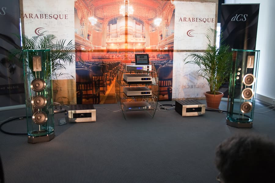 A pair of tall glass-bodied hi-fi loudspeakers on a trade show stand, with hi-fi electronics on a glass rack between them.